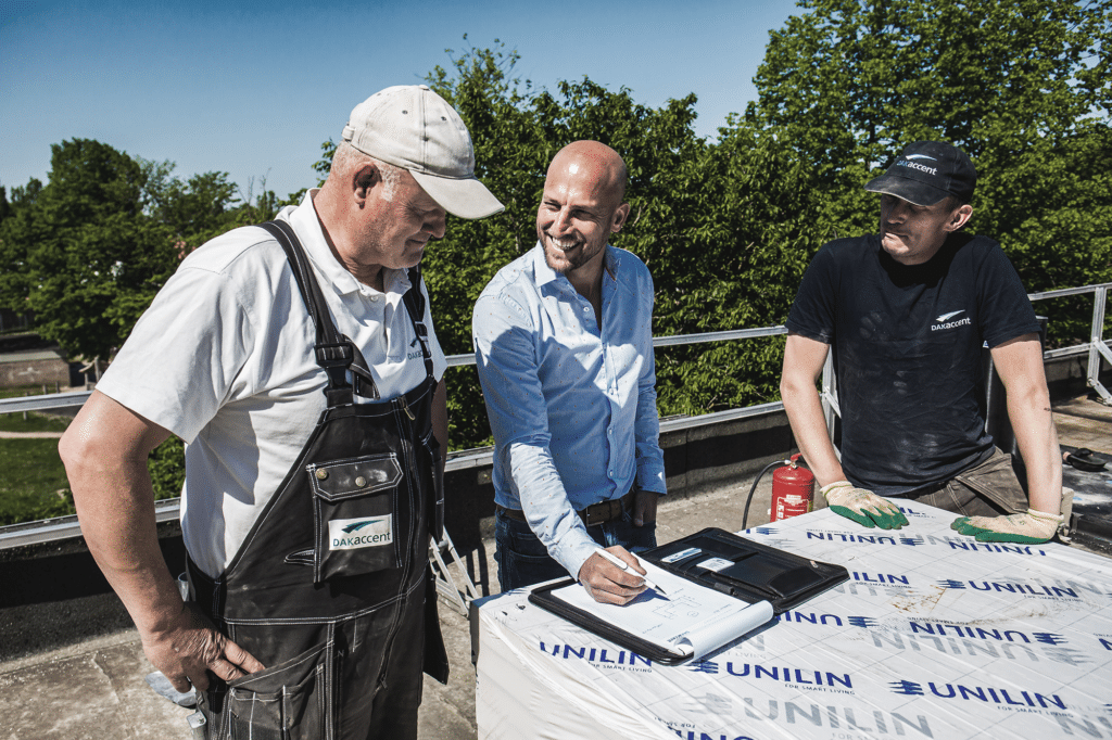 Drie mannen bespreken werkdocumenten op een dak naast isolatiemateriaal.