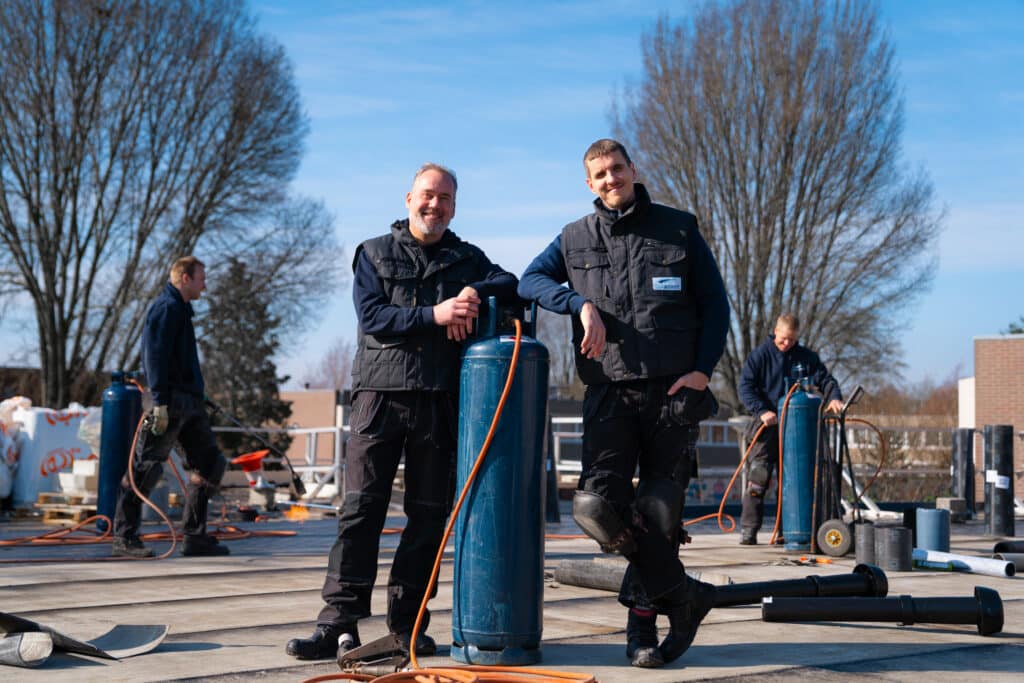 Twee glimlachende mannen in werkuniform leunend tegen een blauwe gasfles op een dak met gereedschap en collega's op de achtergrond.