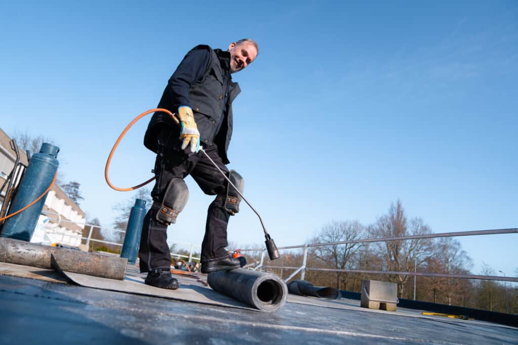 Man in zwarte werkoutfit en beschermende kniepads gebruikt een brander op het dak met gereedschap en een blauwe gasfles op de achtergrond.